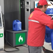 Un atendedor de estación de servicios Shell cargando dos bidones de kerosene en el portamaletas de un auto.